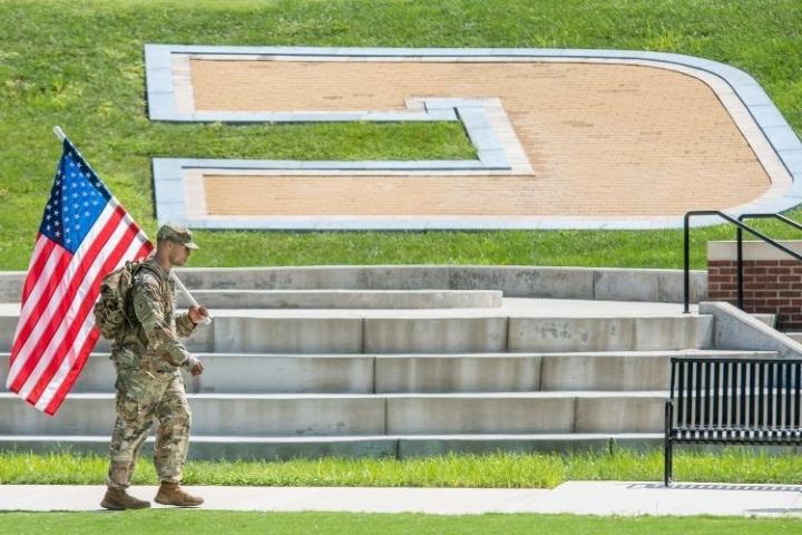 A Solider holding the American Flag walking in front of the Power C on Chamberlain Field.